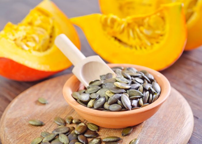 A bowl of pumpkin seeds on a wooden board with fresh cut pumpkin in the background