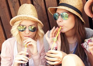 Two women in sunglasses and hats sitting down drinking juices 