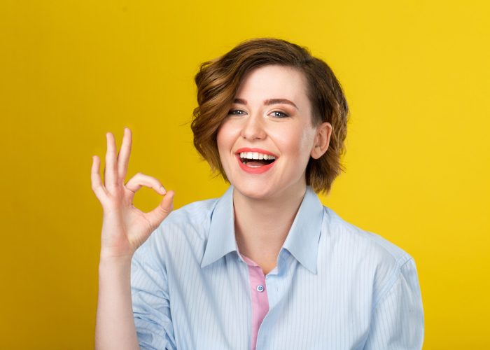 Smiling woman with positive mindset making the ok sign against a yellow background