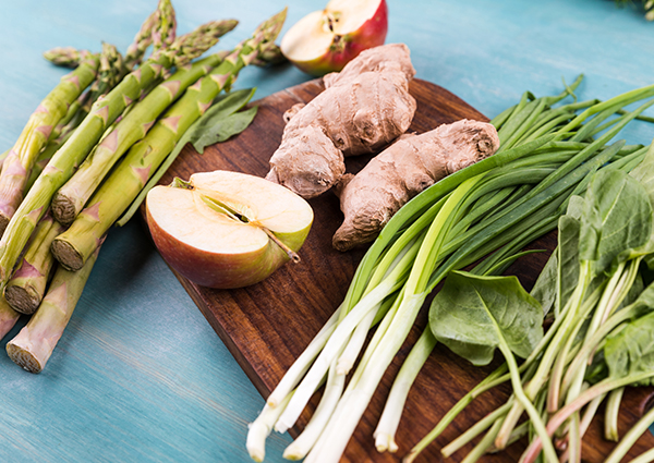 A spread of fresh produce including asparagus, halved apples, spinach, scallions, and fresh ginger on a cutting board