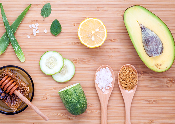 A flat lay of ingredients to make your own DIY face masks at home including half an avocado, aloe vera, fresh honey and honeycomb, cucumber slices, half a lemon, and salt.