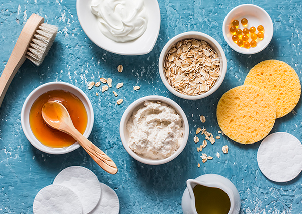 The ingredients for making a homemade oatmeal face make spread out on a blue table including oats, yogurt, and makeup pads