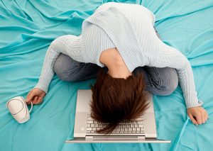 Woman sitting on a bed with her head on her laptop in frustration and exhaustion