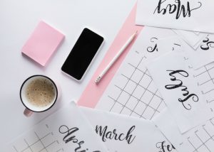 calendar schedule with coffee, a pencil, pink notepads, and a phone on a white table