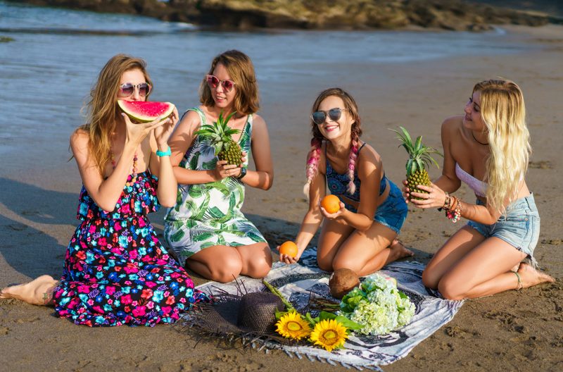 Four women in colourful dresses sitting on the beach enjoying fruits