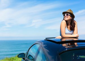 confident woman in a hat and sunglasses leaning out of her car's roof smiling