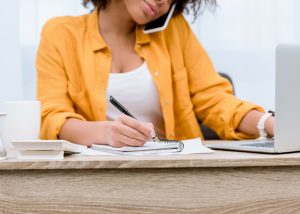 a woman multitasking on the phone, working on her laptop and writing notes 