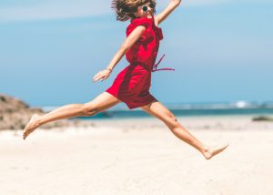 a woman in a red jumpsuit and sunglasses jumping on the beach