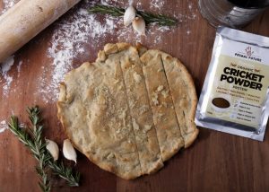 rolled out dough base with sprigs of rosemary, garlic cloves, and a pack of cricket powder on the side