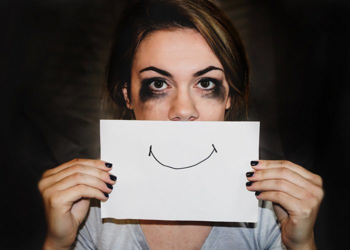 woman with black smudged eyes holding up a piece of paper to her face with a smile drawn on it