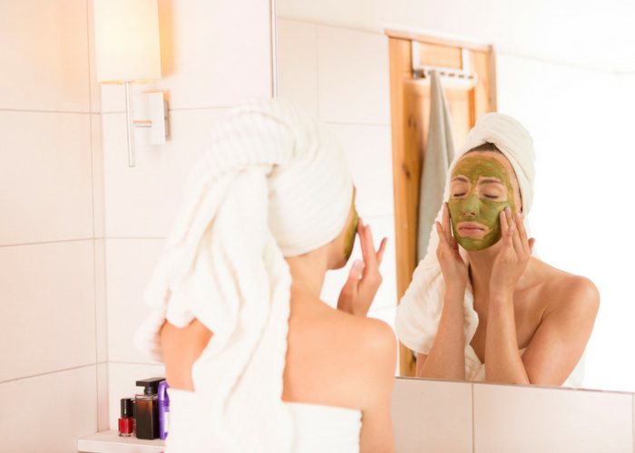 woman with a towel wrapped around her body and head applying a green tea extract face mask as part of her skin care regimen