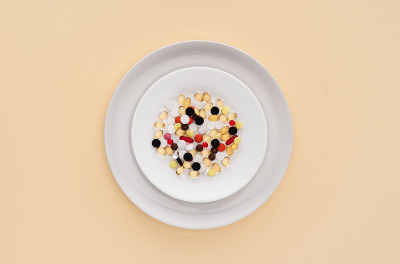 Different colored supplement pills on a white dish, on top of a larger white plate, set against an eggshell yellow background