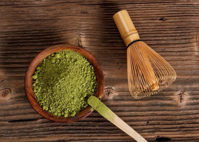 matcha green tea powder in a brown bowl with a wooden scoop and bamboo whisk beside it