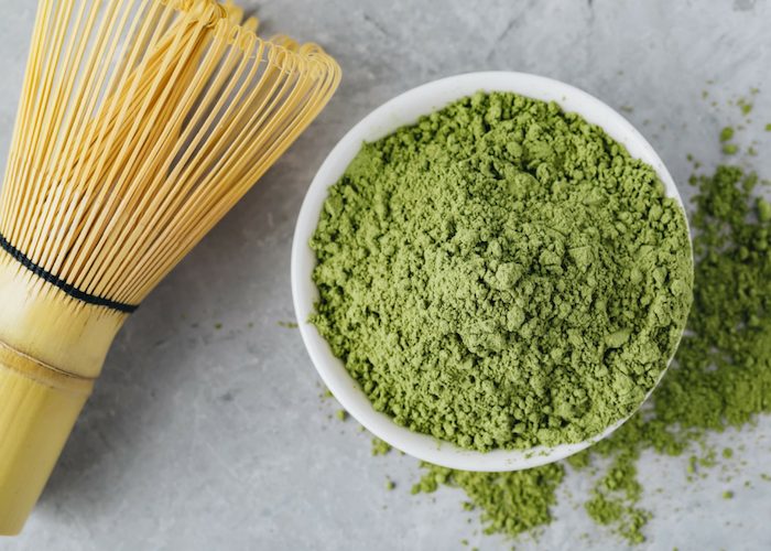 a heaped bowl of matcha green tea powder spilled over onto the table, with a bamboo whisk