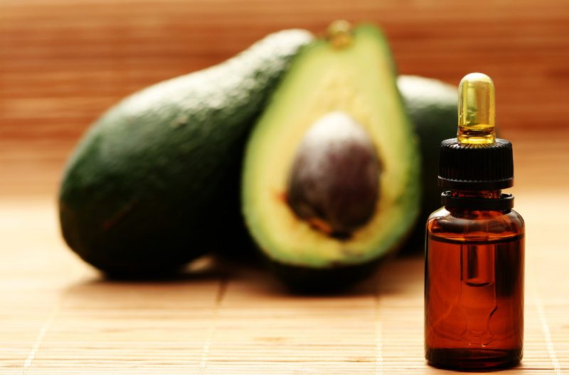 A small dark dropper vial of avocado oil with whole avocados behind it