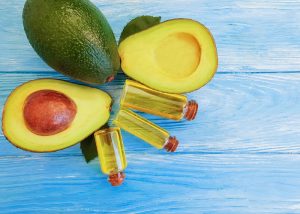 three vials of avocado oil, two halves of an avocado, and a whole avocado on a blue wooden table