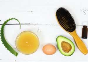 A composition of ingredients needed for healthy hair: fresh aloe vera, avocado oil, an egg, and a hair brush on a white table
