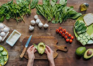 woman with a chopped half of an avocado on a wooden board, and other vegetables around it like garlic, leafy greens, tomatoes