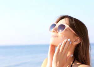 woman with sunglasses at the beach applying sunscreen on her face