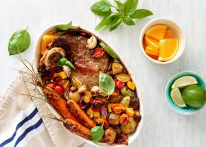 an oven dish filled with meat and roasted vegetables, topped with basil leaves and citrus fruits on the side for zest