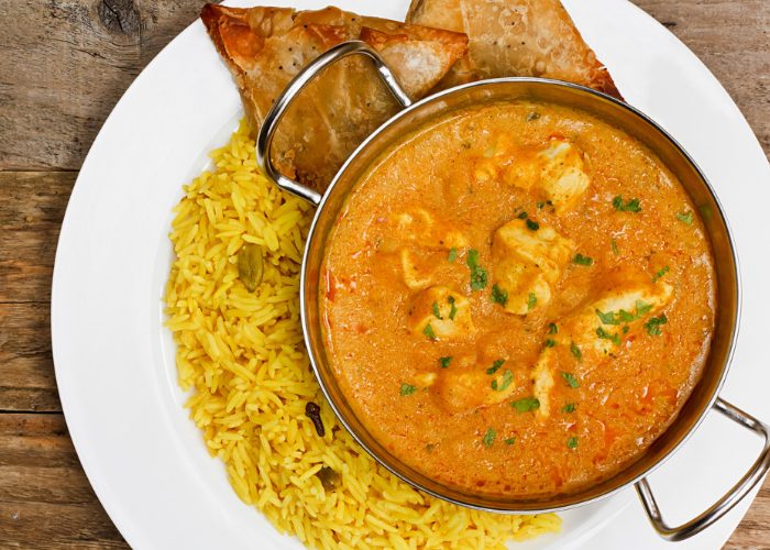 chicken korma curry in a silver dish on a white plate, with sides of yellow rice and samosas