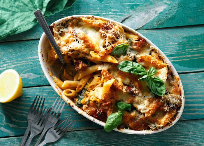 Pasta bake topped with fresh green basil in an oven dish, with four forks and a lemon wedge on a green wooden table