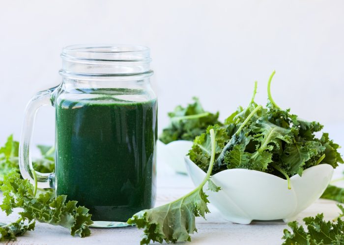 a green mason jar cup of green smoothie with a bowl of kale leaves next to it and kale leaves scattered around