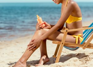 woman in a yellow bikini at the beach, sat on a beach chair, and applying sunscreen on her legs