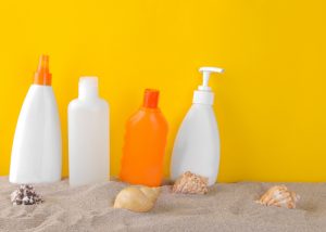 bottles of sunscreen placed in sand against a yellow wall with sea shells scattered around