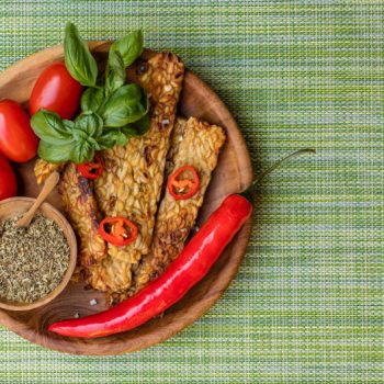 Fried tempeh on a wooden plate with fresh cherry tomatoes, red chili pepper, garnished with basil leaves and cut red chili