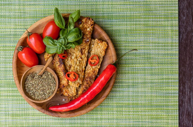 Fried tempeh on a wooden plate with fresh cherry tomatoes, red chili pepper, garnished with basil leaves and cut red chili