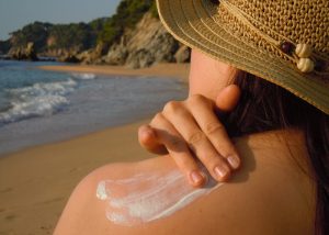 woman with a sun hat at the beach applying sunscreen to her shoulder