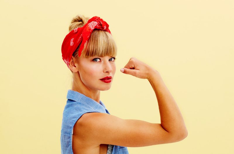 Strong, blonde woman in blue denim sleeveless top and a red headband flexing her biceps against a pale yellow wall, emulating the "We Can Do It!" poster from World War II