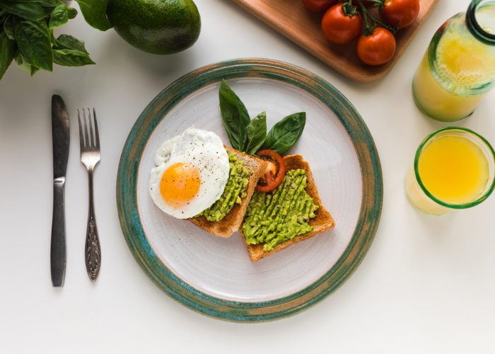 Plant-based breakfast of avocado on toast with a sunny side up egg on a plate, with a fork, knife and glass of orange juice beside it