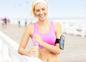 Blonde short-haired woman smiling in light purple sports bra with earphones, an iPhone, and a water bottle after a run