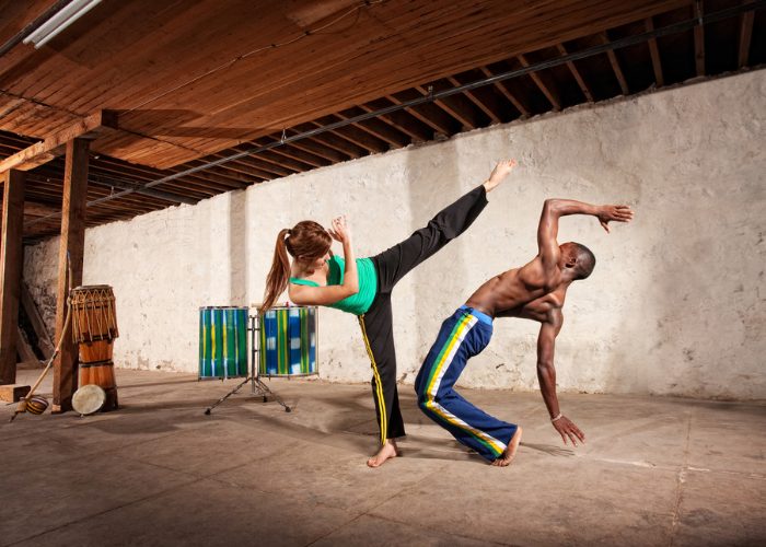 A sparring couple practicing the  Brazilian martial arts, Capoeira. A woman is performing a high kick, and a man is trying to lean back to avoid the kick.
