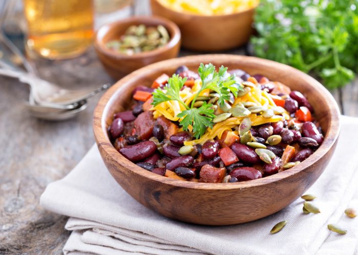 Vegetarian bean chili in a wooden bowl, on a pale tablecloth, on a wooden table with cutlery and ingredients in the background