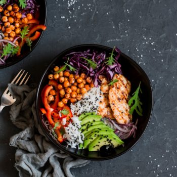 Two bowls of flexitarian salad with grains, vegetables, avocado, some small slices of chicken breast, on a dark grey slate table with two forks beside them