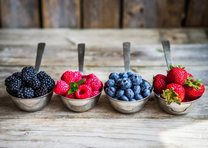 Four different types of berries -  blueberries, raspberries, strawberries, and blackberries - in metal spoons on a wooden table