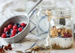 A jar of homemade yogurt with fruits and nuts, next to a metal scoop filled with fresh berries