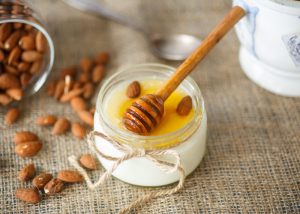 A small jar of homemade yogurt topped with honey and whole almonds, with a honey dipper on the top of the jar, and almonds spilled next to it