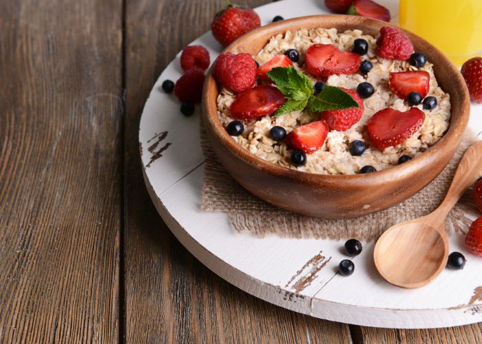 Wooden bowl filled with prebiotic-rich oatmeal, topped with strawberries and blueberries, presented on a white wooden board and wooden spoon