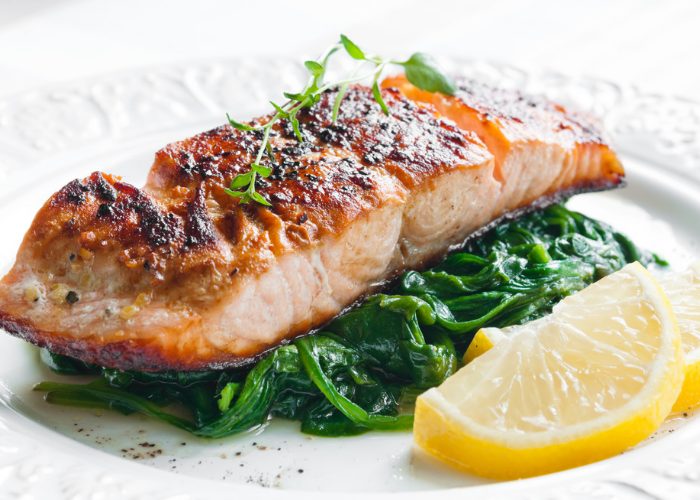 A beautifully presented plate of roast salmon topped with garnish, with a bed of sauteed spinach, and slices of lemon on the side.
