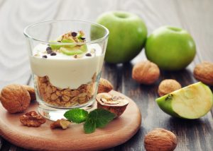 Glass of yogurt with oats, nuts, and green apple, on a wooden dish with walnuts and green apples around it