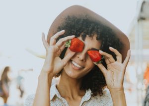 Smiling woman outdoors holding up two strawberries to her eyes