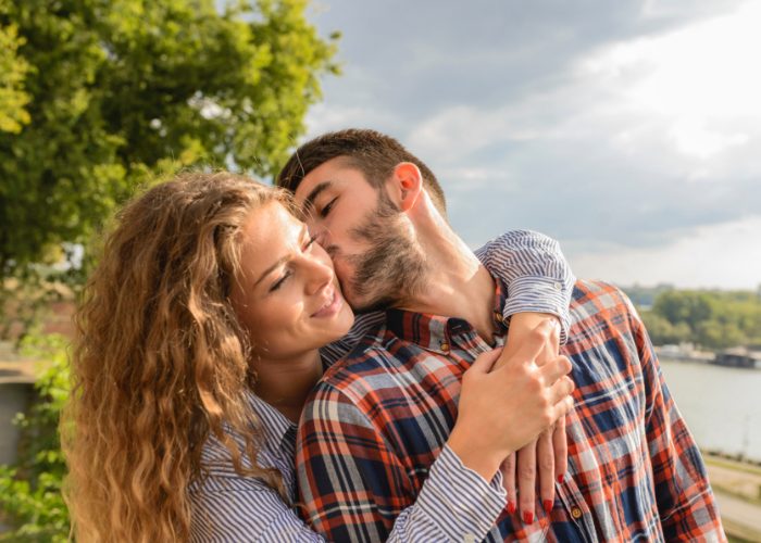 A happy heterosexual couple outdoors, with the man kissing the woman on the cheek as she huge him from behind