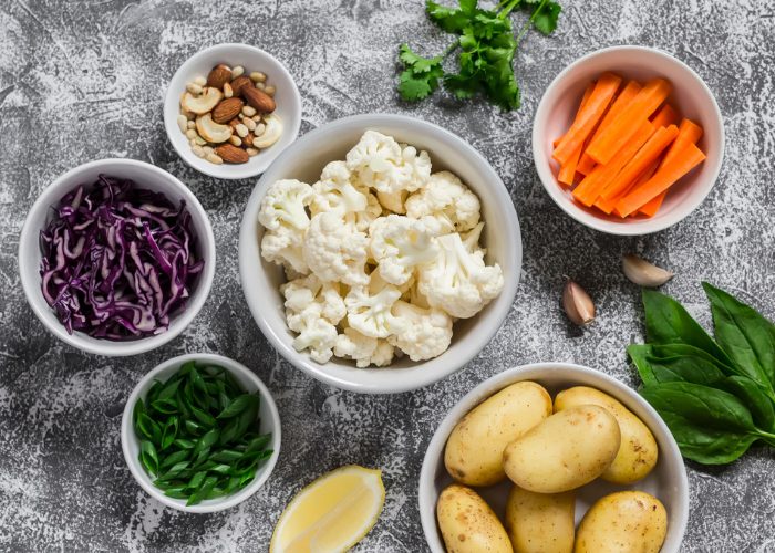 Different bowls with plant-based foods such as cauliflower, potatoes, carrots, nuts and cabbage, on a grey background