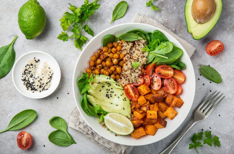 Top down shot of a healthy vegan salad bowl with avocado, sweet potato, spinach, tomatoes, chickpeas, lime, cherry tomatoes, and grains against a grey background with other ingredients laid out neatly