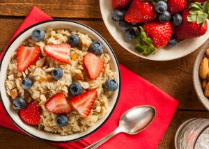 A bowl of oats topped with strawberries and blueberries on a red tablecloth and a spoon set next to it
