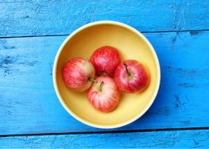 Four whole apples in a yellow bowl on a blue wooden surface